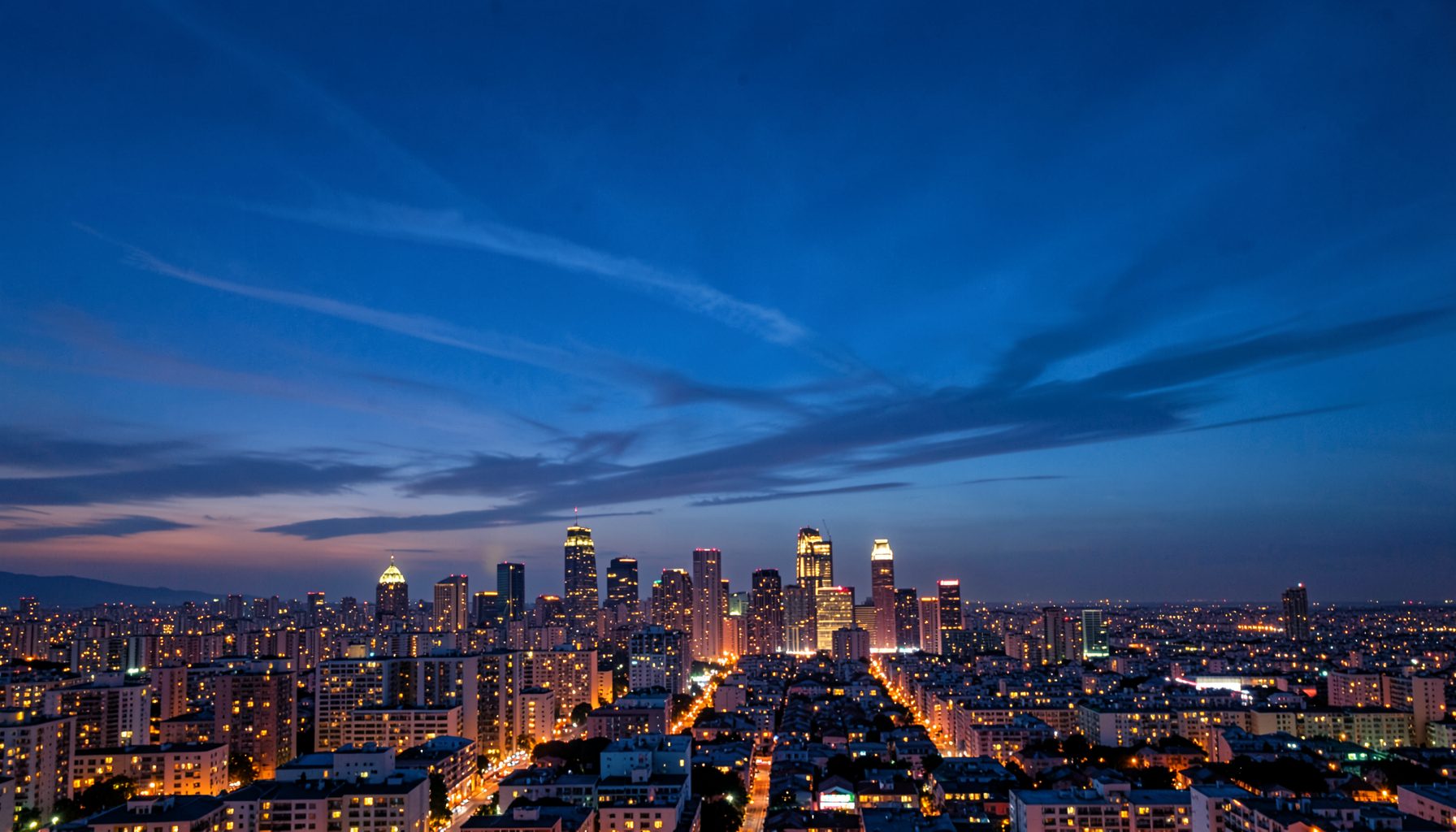 A sweeping time-lapse view of a city skyline at twilight, with warm amber lights glowing against a deep blue sky and soft clouds