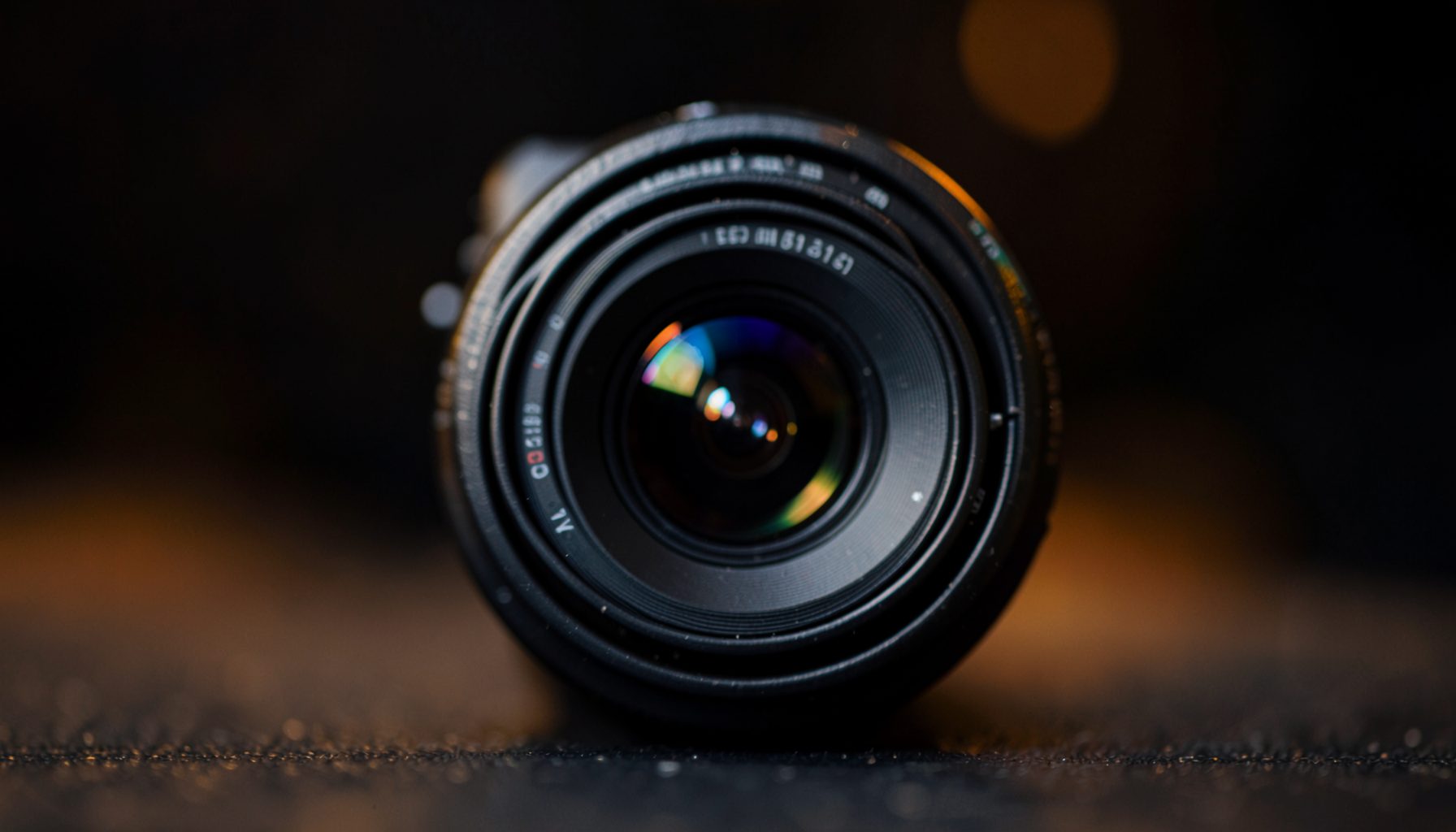A close-up of a compact camera lens with subtle reflections, set against a dark blurred background with warm amber accents
