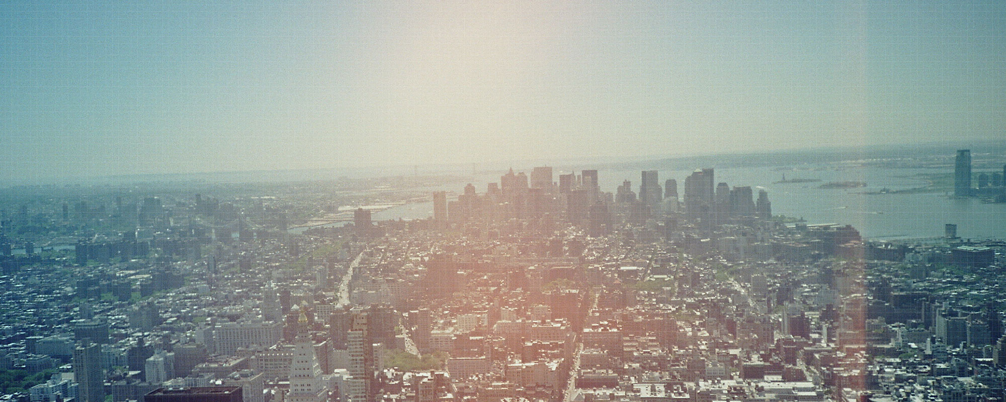 Aerial view of a dense city skyline with skyscrapers and waterfront under hazy sunlight, blue and warm tones