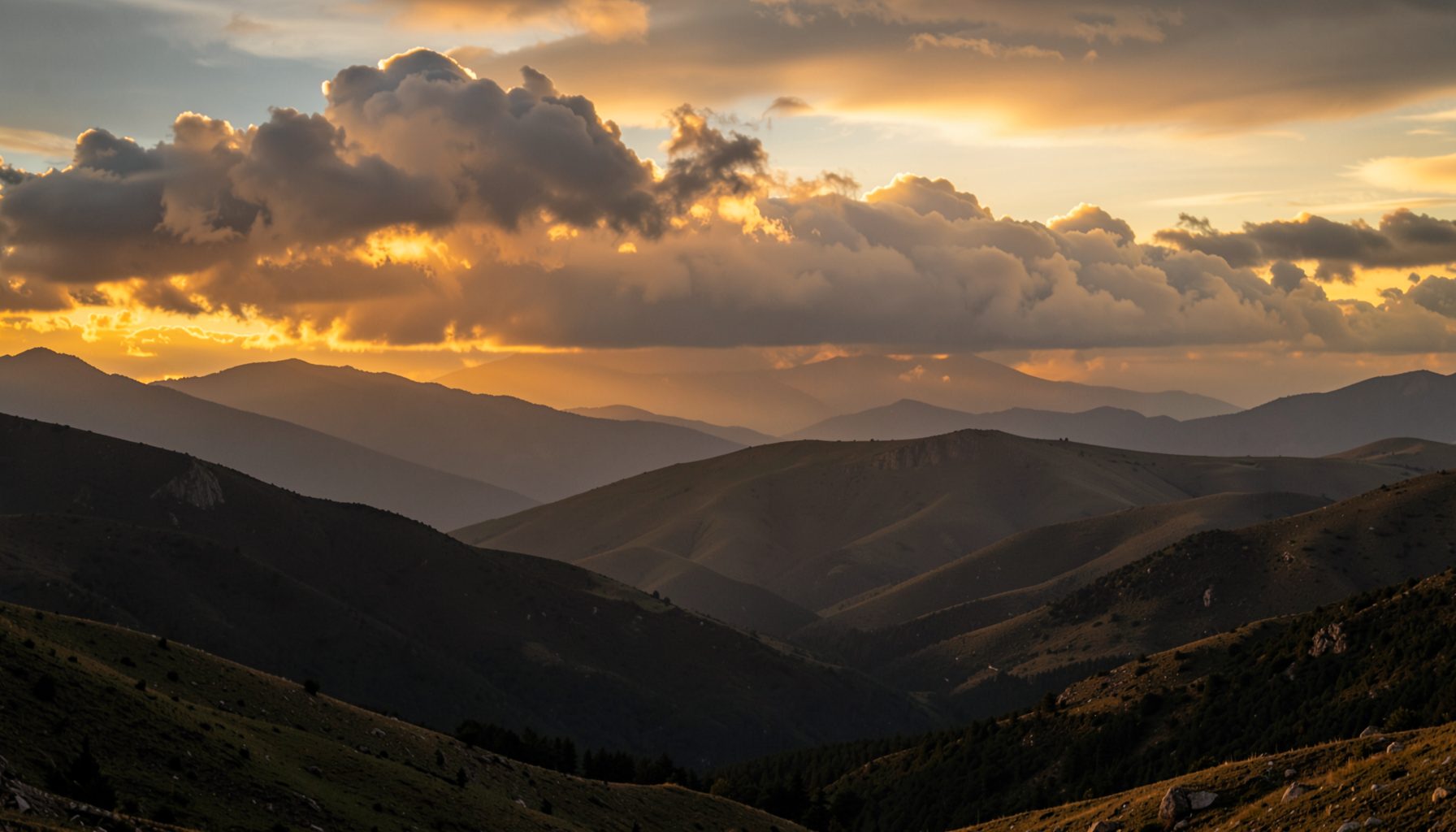 A wide mountain landscape with rolling hills and dramatic clouds at sunset, golden and amber tones