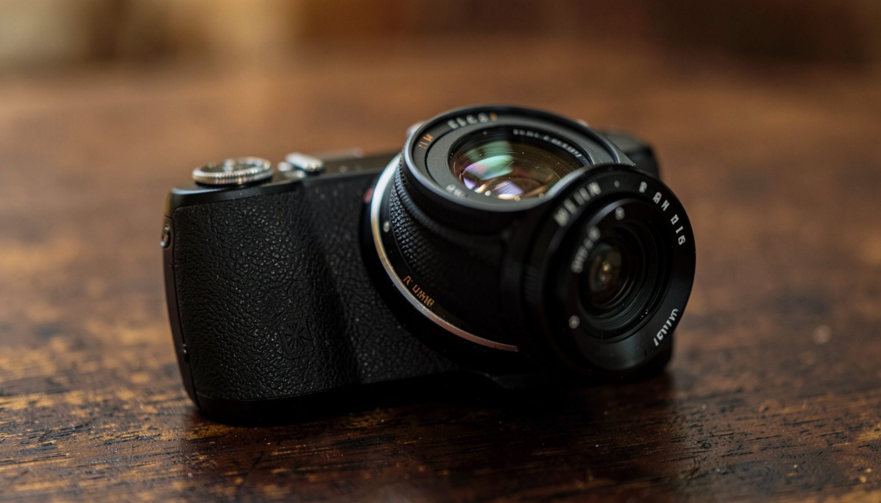 A close-up of a compact camera body with a lens, resting on a dark wooden surface with soft natural light and warm brown tones