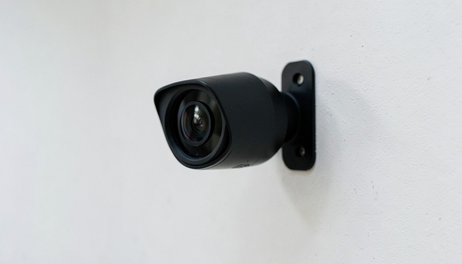 A compact security camera with a dark housing, mounted on a wall bracket under even indoor lighting